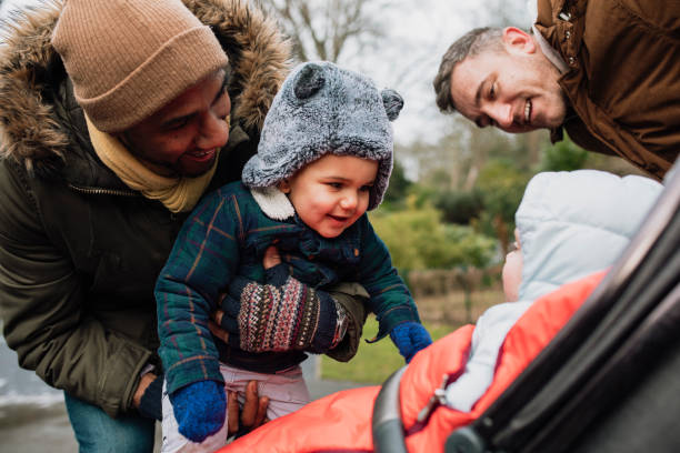 Little Baby Boys Bonding Two Dads are in the park with their baby sons during winter time. They are wearing warm clothing. One of the men is holding his son up close to another baby in a stroller, his son is laughing and smiling at the other baby. baby stroller winter stock pictures, royalty-free photos & images