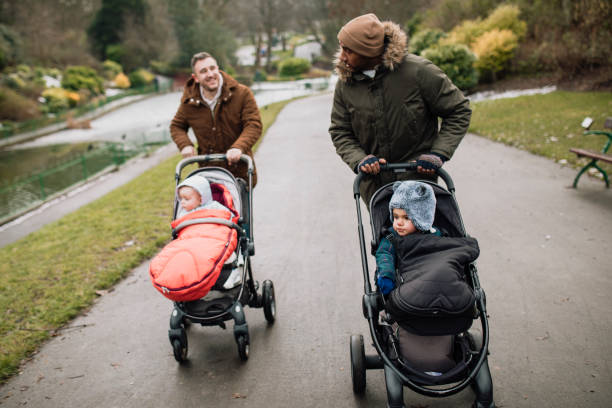 Dads Taking a Walk in the Park with Their Sons Two Dads are walking in the park during winter time. They are wearing warm clothing, pushing their baby sons in strollers and chatting to one another. baby stroller winter stock pictures, royalty-free photos & images