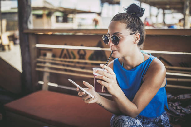 Young woman using mobile phone while drinking juice at beach bar. Young woman enjoying in summer vacation while drinking fruit cocktail and reading text message on cell phone at beach bar. friends in bar with phones stock pictures, royalty-free photos & images