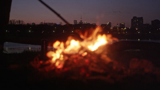 Man and his camp on a riverside. City in background. Making dinner on fire