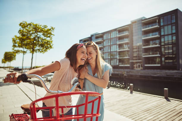 Two young female friends laughing while walking arm in arm together with a bicycle along a city prominade in summer