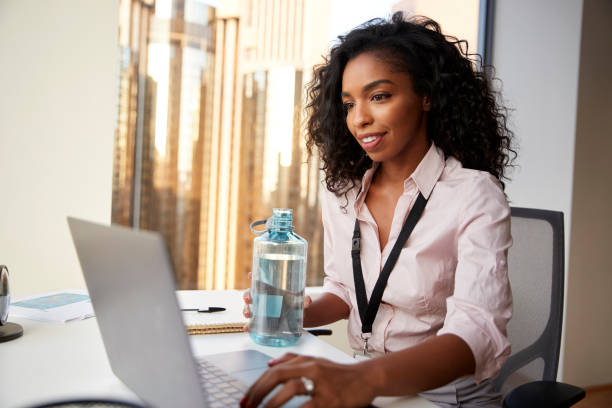businesswoman with laptop sitting at desk keeping hydrated drinking from water bottle in office - water bottle water bottle drinking imagens e fotografias de stock