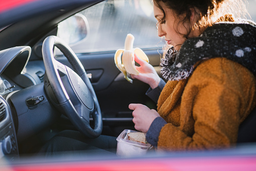 Young businesswoman is sitting in the driver's seat of her car eating a banana.