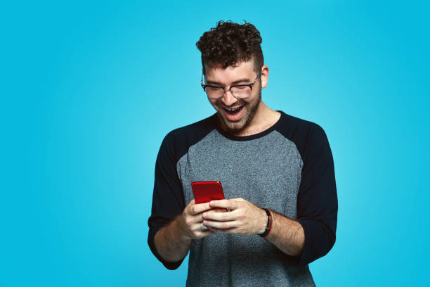 istock Young handsome hipster guy standing against blue wall with copy space, looking at screen of smartphone.d, celebrating 1132298376