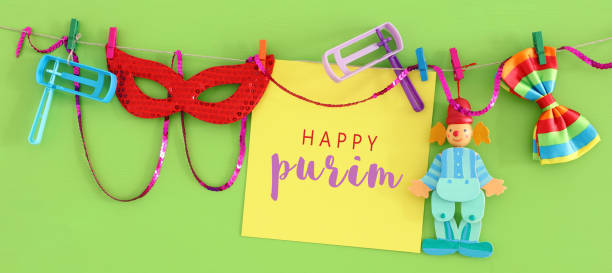 istock Purim celebration concept (jewish carnival holiday) with empty note for text, clown, mask and noisemaker over wooden green background 1132306619
