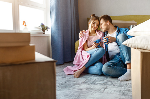 Young family couple bought or rented their first small apartment. Lovely cheerful pople sit on floor and drink from blue cups. Her shoulder covered with pink blanket.