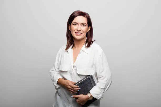 Portrait of mature businesswoman holding diary. Confident female manager is standing against white background. She is having redhead.