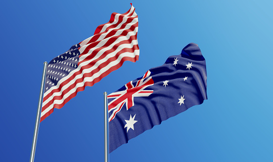 American and Australian flags are waving with wind over  blue sky. Low angle view. Dispute and conflict concept. Horizontal composition with copy space.