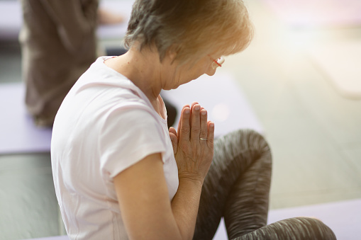 Senior woman sitting on cushion in yoga class, meditating, hands folded in front of chest (prayer pose)
