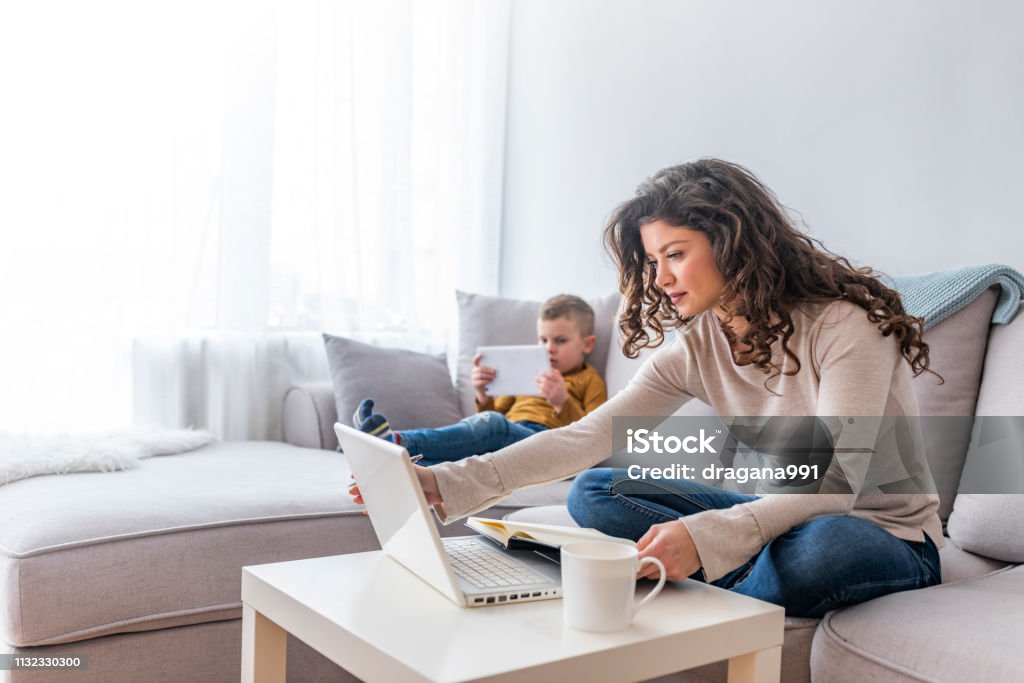 Busy Mother Working From Home With son Mother working on laptop at home, while her child using tablet. Boy and mother using tablet and laptop. Modern family. Busy Mother Working From Home With son Working At Home Stock Photo