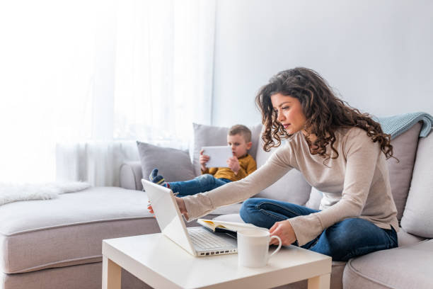 Busy Mother Working From Home With son Mother working on laptop at home, while her child using tablet. Boy and mother using tablet and laptop. Modern family. Busy Mother Working From Home With son working at home with children stock pictures, royalty-free photos & images