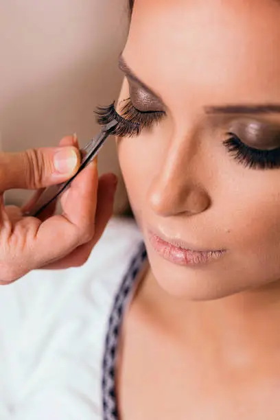 Make up artist working in a make up studio, putting artificial eyelashes on female client's eyelids with a tweezers