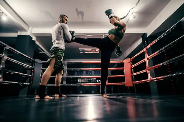 Photo of Powerful caucasian boxer girl in sportswear, with bandage on feet and boxing gloves on hands practicing with trainer in ring.