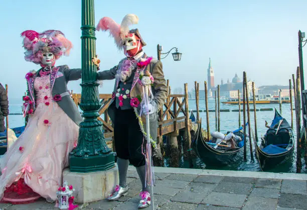 Models disguised in venetian carnival costumes are posing near some docked gondolas, both are touching à street light pylon and watching at the camera in the background it can be seen the San Giorgio Maggiore church,she is wearing a beautiful pink dress,beautifully decorated with red roses,a gorgeous hat dight with feathers and flowers,he is wearing a jacket dight with roses ,silver mask and feathers on the top of the head,she is holding a decorated mirror and he is holding a baston also decorated with flowers.