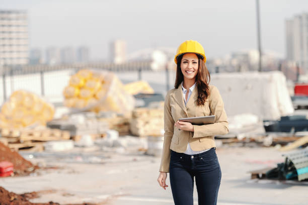 piękna kaukaska kobieta architekt z długimi brązowymi włosami, zębaty uśmiech i kask na głowie trzymając tabletki podczas spaceru na placu budowy. - architect female women construction zdjęcia i obrazy z banku zdjęć