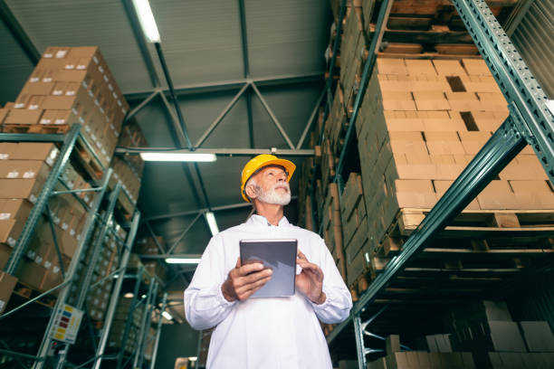 istock Bottom view of gray bearded worker in white uniform and protective helmet on head holding tablet and looking up while standing in warehouse. 1132357680