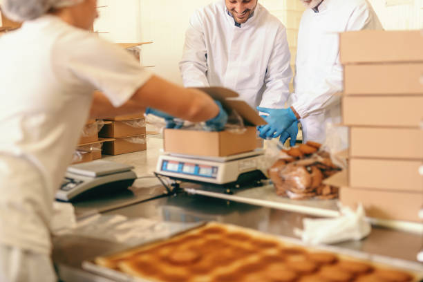 istock Three workers in sterile uniforms, rubber gloves and hear nets on heads packing cookies in boxes. Food plant interior. 1132358908