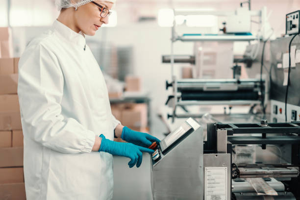 Young female employee in sterile uniform and blue rubber gloves turning on packing machine while standing in food factory. Young female employee in sterile uniform and blue rubber gloves turning on packing machine while standing in food factory. food and drink industry photos stock pictures, royalty-free photos & images