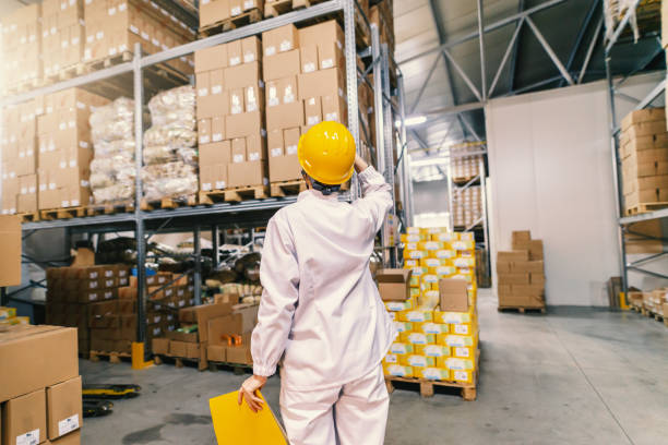 istock Young Caucasian employee in white uniform, protective helmet and with folder in hand looking up and standing in warehouse. Backs turned. 1132360437