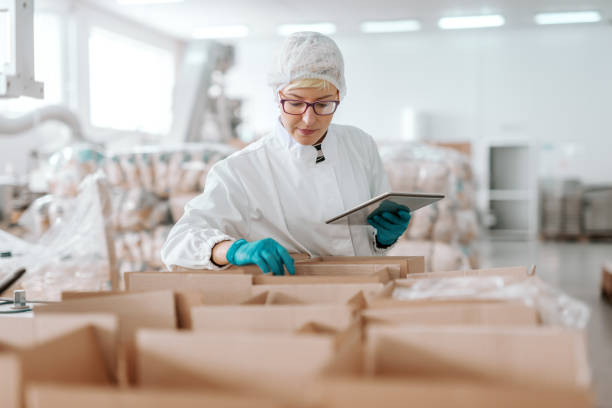 istock Young Caucasian employee in sterile uniform holding tablet and counting products in boxes. Food factory interior. 1132361078