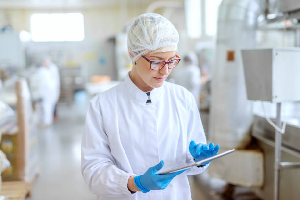 supervisor en uniforme estéril y con anteojos usando tableta para controlar el flujo de trabajo en la fábrica de alimentos. - food hygiene fotografías e imágenes de stock