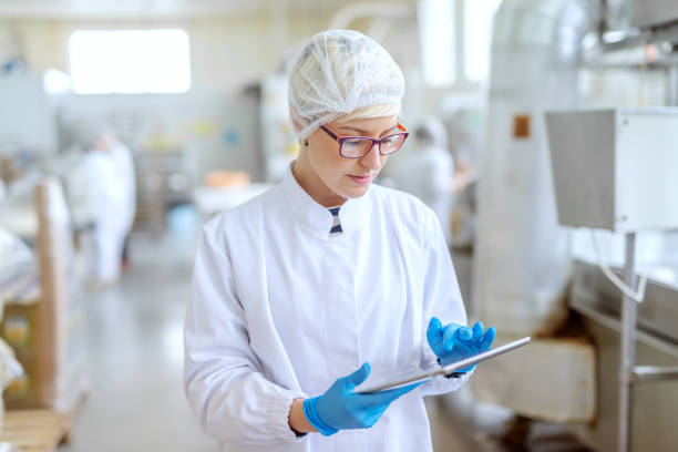 supervisor in sterile uniform and with eyeglasses using tablet for controlling workflow in food factory. - food hygiene imagens e fotografias de stock