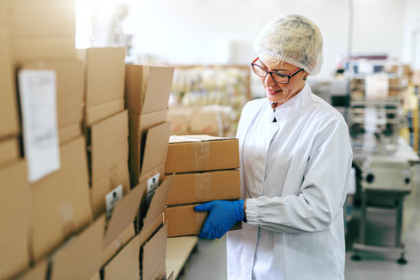 istock Smiling female blonde employee in sterile uniform and with eyeglasses putting boxes on stack. Food factory interior. 1132361531