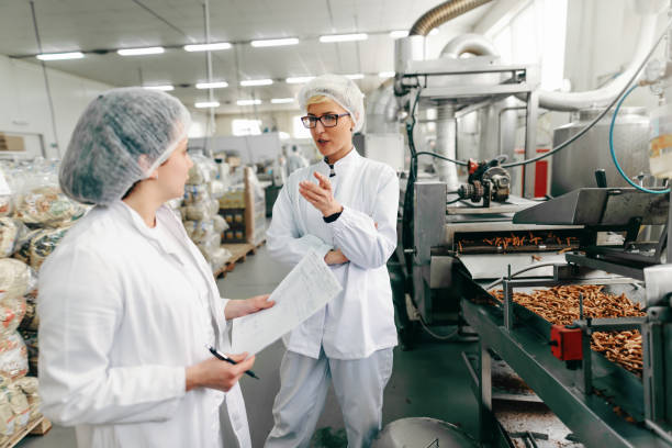 coworkers in white uniforms and with sterile caps on heads discussing about quality of products while standing in food factory. - food and drink industry imagens e fotografias de stock