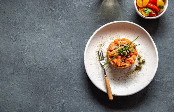 SALMON TARTAR with capers and purple onion on white plate, gray background.