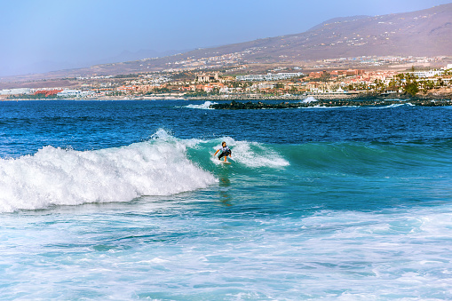Costa Adeje, Spain - February 8, 2019: Surfer girl on a ocean wave against the background of a popular resort in the southern coast of the Canary island of Tenerife and the mountains in kalima haze.