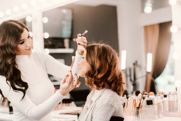Sophisticated beautiful caucasian brunette sitting in beauty salon while makeup artist putting blush on woman's cheeks. Sophisticated beautiful caucasian brunette sitting in beauty salon while makeup artist putting blush on woman's cheeks. makeup artist stock pictures, royalty-free photos & images