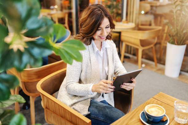 istock Gorgeous smiling brunette dressed casual using tablet and sitting in cafeteria. On desk coffee and glass of water. 1132385101