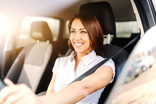 Portrait of beautiful caucasian woman with toothy smile and brown hair driving car. Hand on steering wheel.