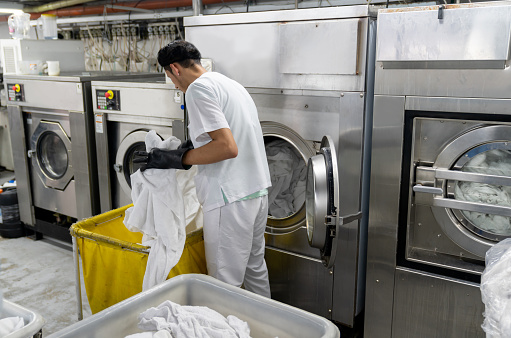 Latin american man loading washing machine with dirty bed sheets at an industrial laundry service wearing protective gloves