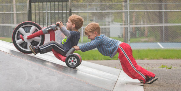 Children Using Cooperation and Teamwork to Reach the Top of a Skatepark Ramp A young boy riding a "Big Wheel" is unable to reach the top of a skatepark ramp until a friend lends a hand and together they reach their goal tricycle stock pictures, royalty-free photos & images