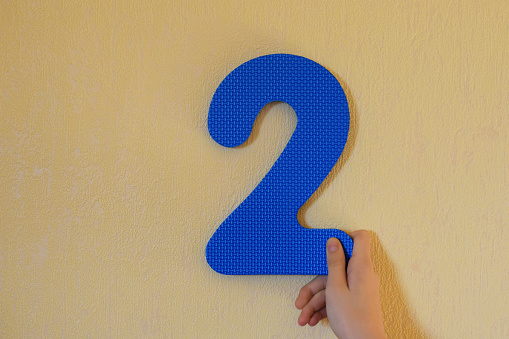 Hand holding blue number two on yellow wall background with copy space for text. Second anniversary birthday design or educational children toys for learning colors and numbers concept.