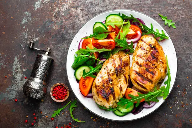 Grilled chicken breast. Fried chicken fillet and fresh vegetable salad of tomatoes, cucumbers and arugula leaves. Chicken meat with salad. Healthy food. Flat lay. Top view. Dark background