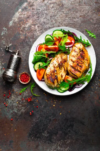 Grilled chicken breast. Fried chicken fillet and fresh vegetable salad of tomatoes, cucumbers and arugula leaves. Chicken meat with salad. Healthy food. Flat lay. Top view. Dark background