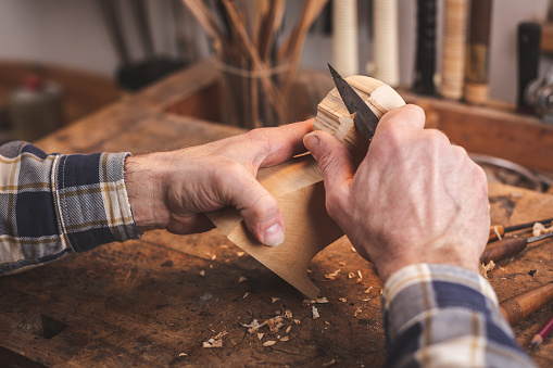 Hands of a man using a knife to carve a small piece of wood on a rustic workbench.