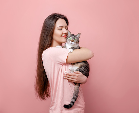 cat licks girl nose, young attractive woman hugging cat in hands, pink background