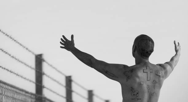 Guy with doodle tattoos with arms reached out at barbed wire fence. stock photo