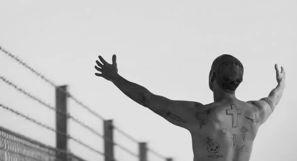 Photo of Guy with doodle tattoos with arms reached out at barbed wire fence.