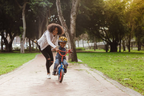Mom teaching her son biking at park Cute boy learning to ride a bicycle with his mother. Woman teaching son to ride bicycle at park. weekend activities photos stock pictures, royalty-free photos & images