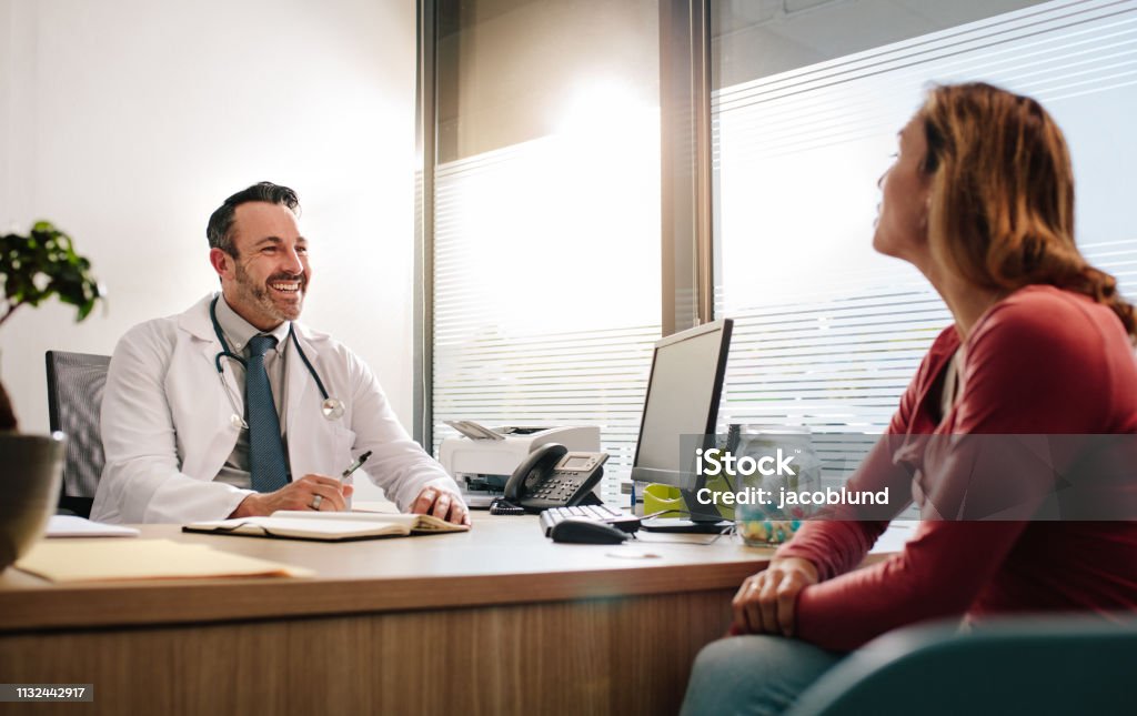 Friendly doctor consulting his patient Doctor talking with female patient sitting across the desk in clinic. Mature male doctor in white medical coat consulting his patient. Doctor Stock Photo