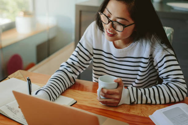 Asian female student working on her assignment Woman making notes in her dairy holding a cup of coffee and laptop in front on table. Asian female student working on her assignment at home. asian adult student stock pictures, royalty-free photos & images