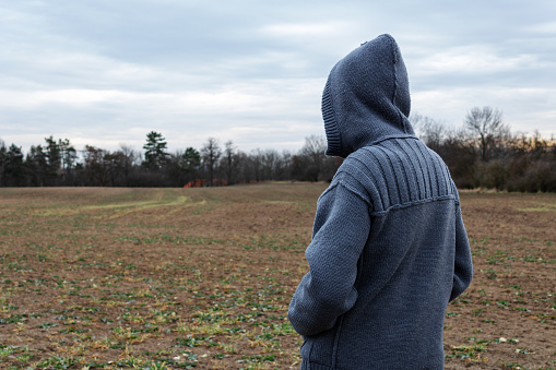 Conceptual anonymous unidentified person in hood is standing on field and looking at property in background
