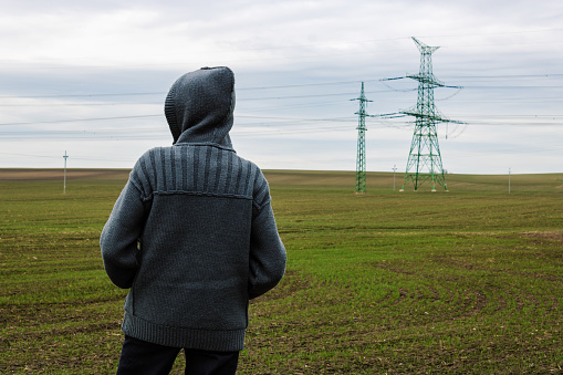 Conceptual anonymous unidentified person in hood is standing on field in front of electric pylon and looking