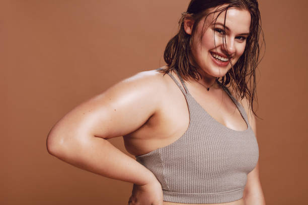 Plus size female model in sports bra Plus size female model in sports bra looking at camera and smiling. Positive oversized young caucasian woman against brown background. sports bra stock pictures, royalty-free photos & images