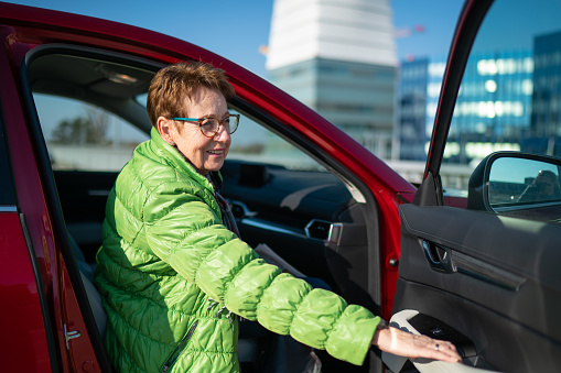 happy smiling senior woman 70 years old get in to the car closing the door of passenger side on sunny winter day on parking space, shallow focus