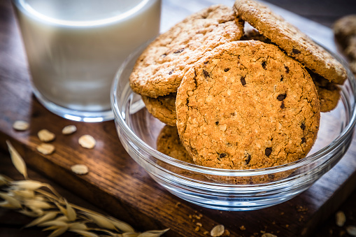 Homemade oatmeal cookies with chocolate chips on a crystal bowl upon a cutting board on rustic wooden table. Alongside the bowl is a glass full of milk.. Main focus is on cookies. Predominant color is brown. Low key DSLR photo taken with Canon EOS 6D Mark II and Canon EF 24-105 mm f/4L